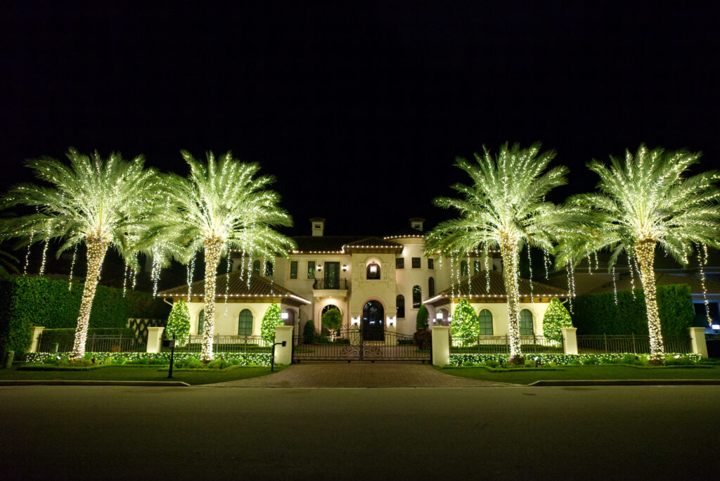 "Four majestic Medjool palm trees, beautifully illuminated by warm white holiday lights wrapped around their trunks and extending up to the fronds, feature additional long string lights hanging from each frond, creating a stunning, majestic ambiance in front of a lovely home during the festive season."